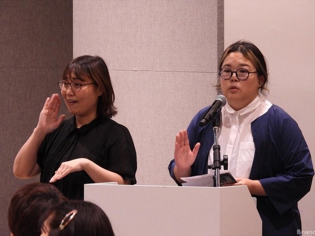 白い演台に立つ2人の人物。右側の人物はマイクに向かって話し、左側の人物は手話を行っている