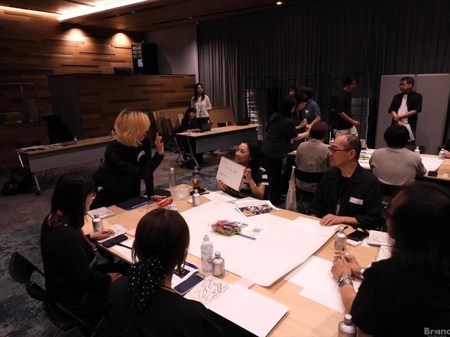 複数の人が大きなテーブルを囲んで座っているワークショップの様子。小さなホワイトボードを掲げる女性や、立って手を振る人物など、参加者たちが和やかに交流している