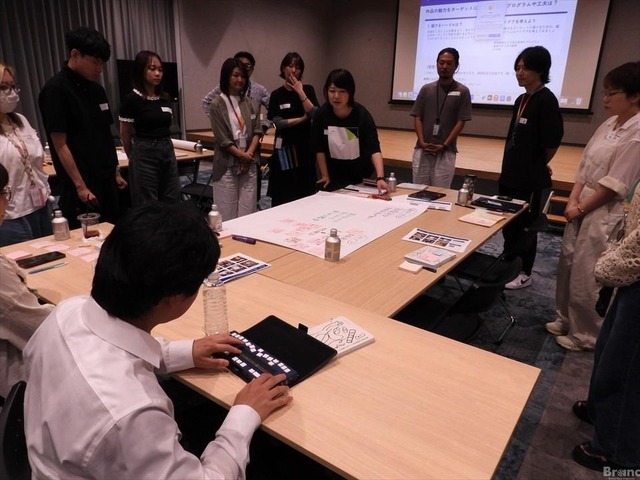 大きなテーブルを囲んで意見交換をするワークショップの様子。手前には点字ディスプレイを操作している白いシャツの男性が座り、奥では複数の参加者が立って模造紙を囲みながら、黒い服の女性の説明を聞いている